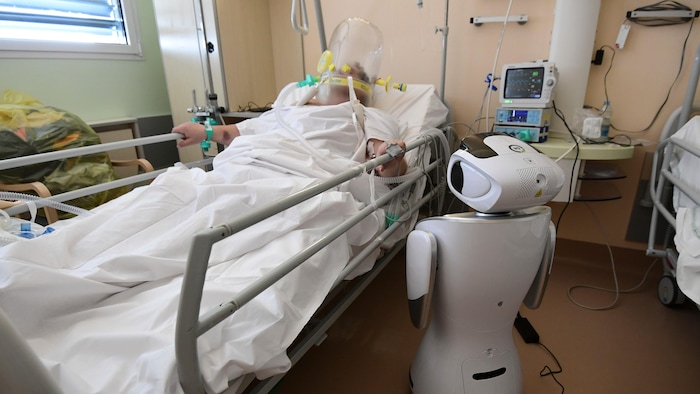 Un robot blanc se tient à la droite d'un lit d'hôpital où un patient est alité. 