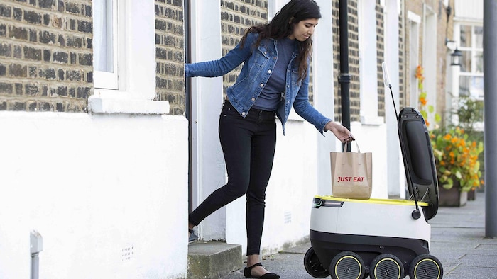 Une femme, devant le pas de sa porte de maison, ramasse dans le petit coffre d'un robot un sac contenant la nourriture qu'elle a commandée.