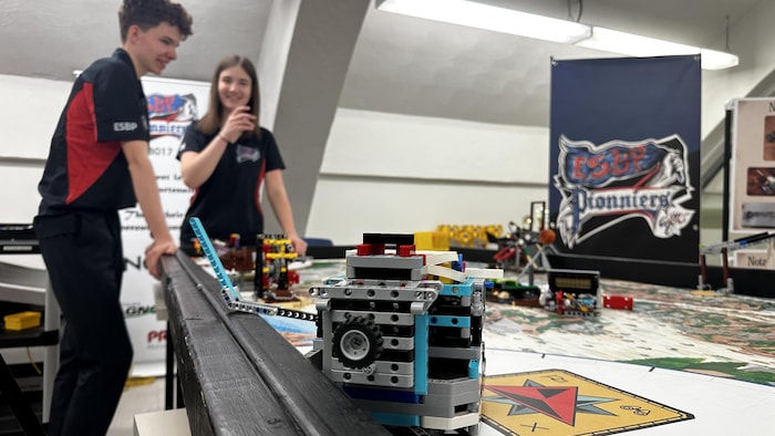 Un robot en légo est sur une table de jeu au premier plan, deux élèves rient en arrière. 