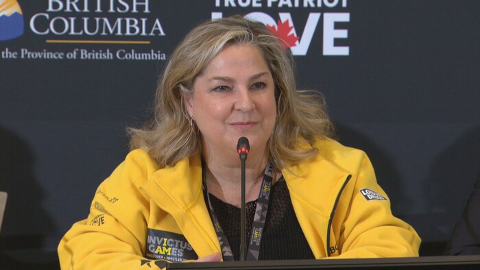 Une femme a un micro devant elle. Elle est assise et porte une veste où il est écrit « Jeux Invictus », en anglais.