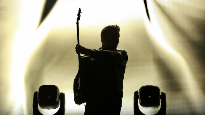 L'ombre du chanteur Roch Voisine, sa guitare levée d'un côté, se détache de l'éclairage en arrière-scène.
