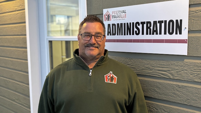 Rock Boudreau sourit chaleureusement devant le bureau administratif du festival, arborant fièrement le logo de l'événement sur sa veste.
