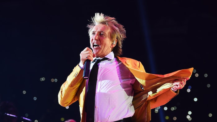 Un homme vêtu d'un veston orange chante dans un micro sur scène.