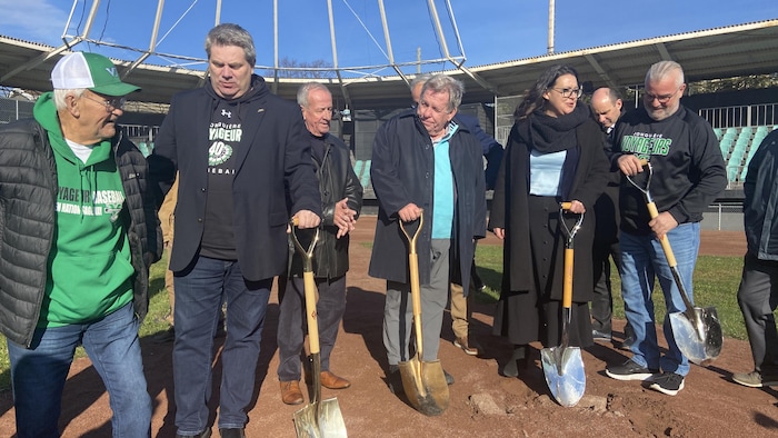 Plusieurs personnes participent à une pelletée de terre officielle sur un terrain de baseball. Certains portent des vêtements aux couleurs des Voyageurs de Jonquière. Quatre pelles dorées sont plantées dans le sol rouge devant un stade sous un ciel bleu.