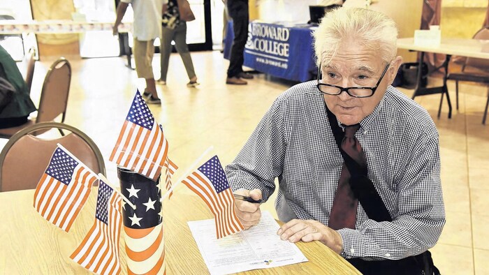 Rodney Nichols assis à un bureau remplit un document. Sur le bureau se trouvent des drapeaux américains.