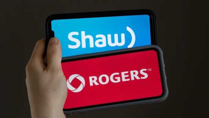 Une main tient deux cellulaires, l'un affichant le logo de Shaw, l'autre le logo de Rogers.