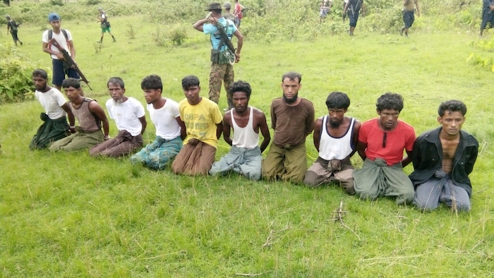 Dix hommes à genoux dans un champ avec les mains derrière le dos, entourés d'hommes armés.