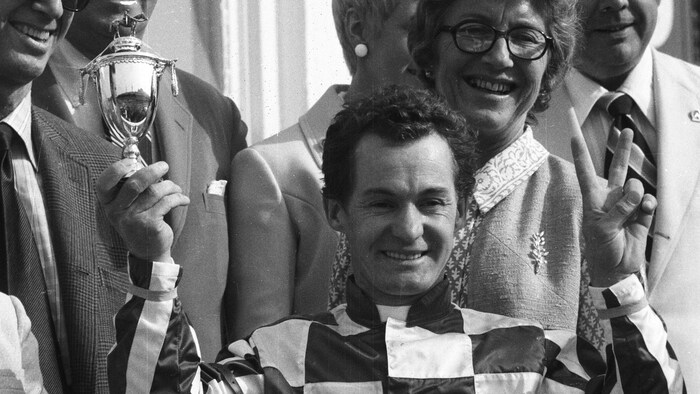 Photo en noir et blanc d'un jockey vêtu d'une chemise à carreaux, entouré de dignitaires. Il fait un signe de victoire des doigts de la main gauche et tient un petit trophée dans la main droite.