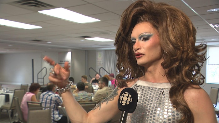 Une drag queen en entrevue, micro pointé vers elle, dans une salle de conférence d'un hôtel.