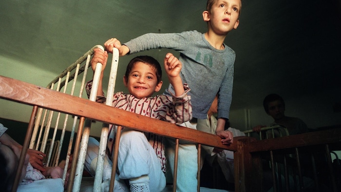 Deux enfants sont dans un orphelinat et se tiennent derrière des barreaux de bois. Un enfant sourit à la caméra et l'autre regarde au loin.