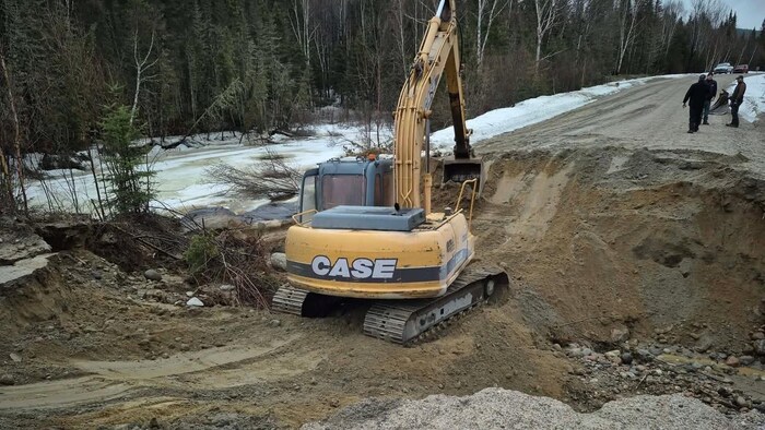 Une excavatrice jaune intervient sur une route de terre gravement endommagée par une crevasse. La scène se déroule en bordure d'une forêt enneigée et d'un cours d'eau, sous l'œil de trois travailleurs.
