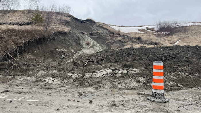 Un glissement de terrain où un amas de terre argileuse a été retourné. Un cône orange est placé devant, en bordure de la route. 