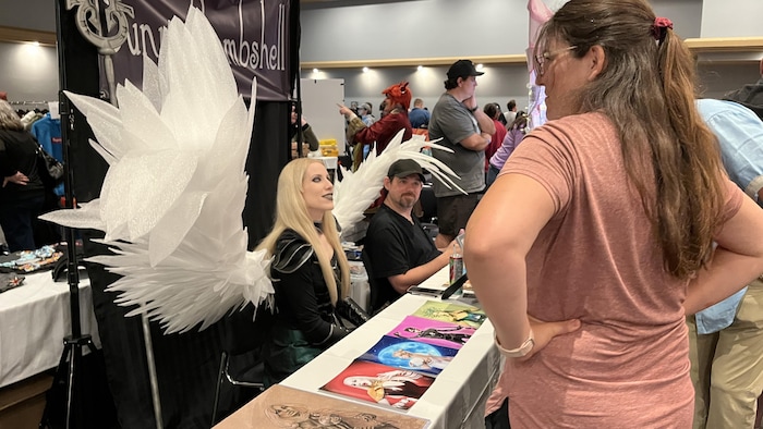 Une femme portant un costume tiré d'un jeu vidéo est assise derrière un kiosque d'exposant au RouynCon.