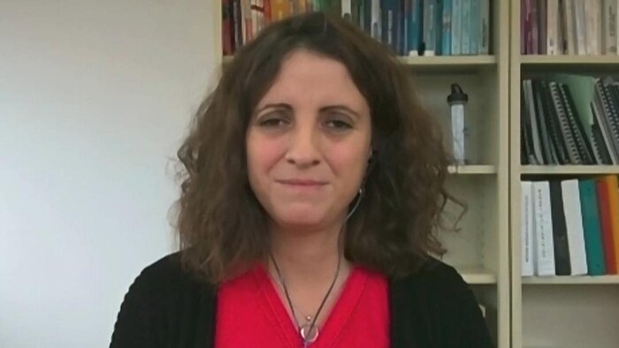 البروفيسورة روكسان بورجيس دا سيلفا تعطي مقابلة بواسطة الكاميرا ونرى خلفها مكتبة ملأى بالكتب.