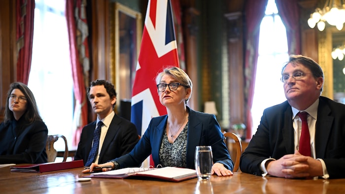 Yvette Cooper est assise en salle de conférence avec deux hommes et une autre femme, devant un drapeau du Royaume-Uni.