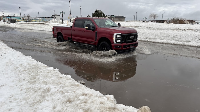 Une camionnette rouge circule dans une large flaque d'eau sur une route bordée de hauts bancs de neige sale