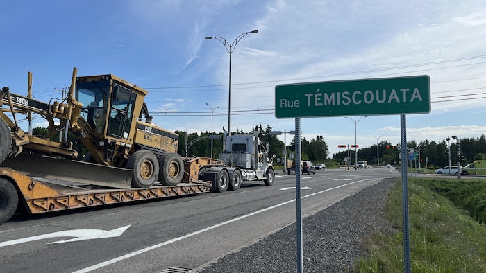 Un camion-remorque à 10 roues transporte un tracteur et est arrêté au feu rouge. Une affiche en avant plan indique "rue Témiscouata"