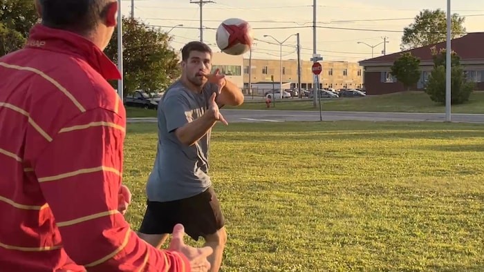 Deux hommes sur un terrain d'herbe. L'un d'eux lance un ballon de rugby blanc et rouge.