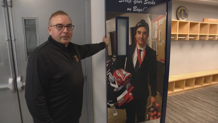 Russ Herold se tient à côté d'une affiche représentant son fils, Adam, dans les vestiaires de l'équipe des Pat Canadians de Regina.
