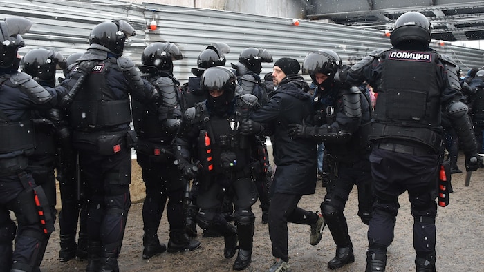 Des dizaines de policiers antiémeute sont en ligne devant un panneau de métal. Un homme est emmené par deux policiers.