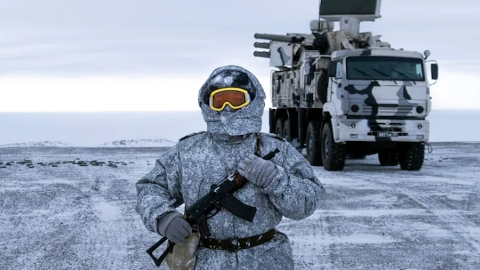 Un soldat emmitouflé dans un habit de neige et portant des lunettes de ski tient une arme dans ses mains, et marche dans le froid devant un immense véhicule militaire.