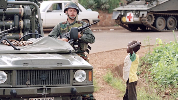 Next to a military vehicle, an armed soldier stands next to a child with his hands on his head.