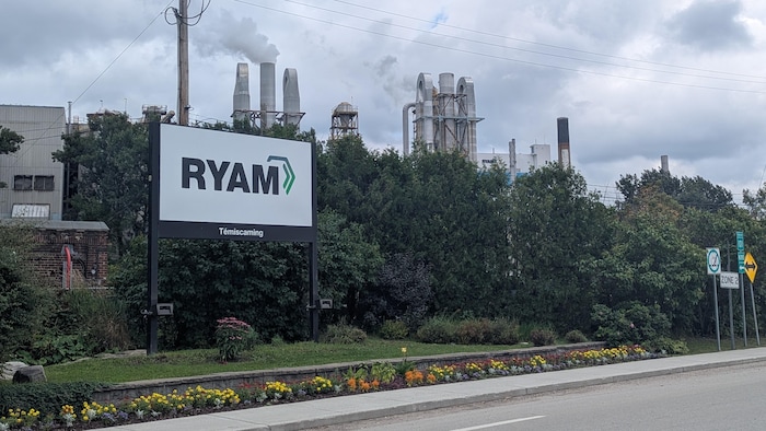 L'affiche de RYAM à l'avant-plan et une partie de l'usine derrière des arbres.