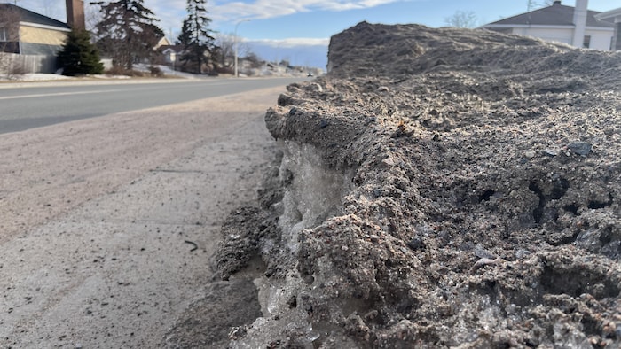 Un amas de neige sale et de terre borde une rue résidentielle sous un ciel bleu dégagé.