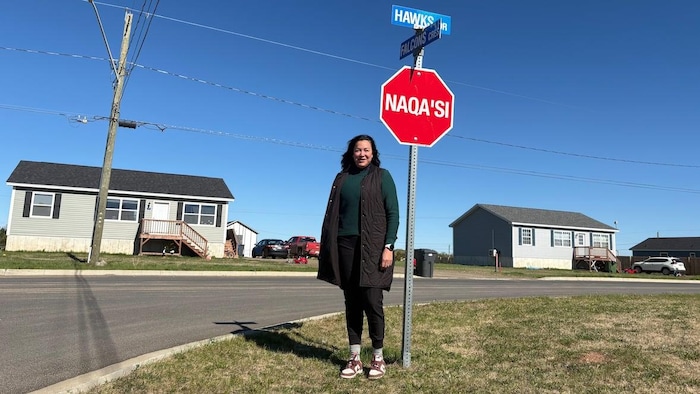 Une femme autochtone au coin d'une rue dans un quartier résidentiel. À sa droite, un panneau d'arrêt écrit en langue mi'kmaq.