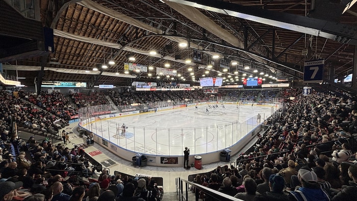 Une vue en plongée montre un match de hockey dans une aréna bondée. Les joueurs évoluent sur la glace entourée de bandes publicitaires. La structure se distingue par un imposant plafond en bois soutenu par des poutres métalliques sombres.