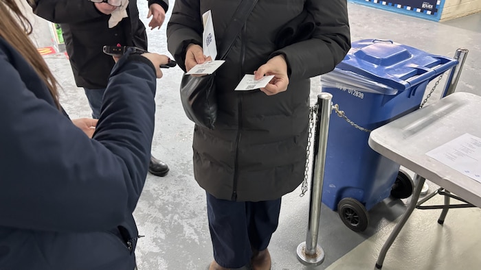 Une personne en manteau d'hiver présente deux billets papier munis de codes QR à un employé tenant un scanner portatif. La scène se déroule à une entrée intérieure, à côté d'une borne de délimitation et d'un bac de recyclage bleu.