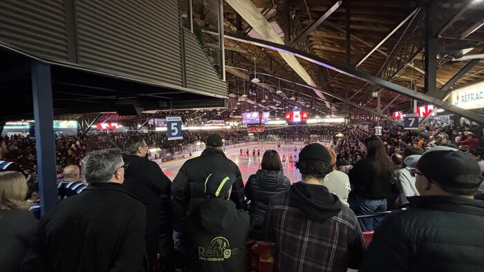 Cette photo montre l'intérieur d'un aréna de hockey bondé, capturée depuis les gradins derrière un groupe de spectateurs. Au centre, les joueurs sont sur la patinoire éclairée sous une imposante charpente en bois et des poutres métalliques sombres.