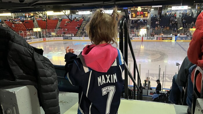 De dos, une jeune fille aux cheveux ébouriffés porte un chandail de hockey bleu marqué du nom "MAXIM" et du numéro 7. Elle observe une patinoire illuminée depuis les gradins d'un aréna où des sièges rouges sont visibles.