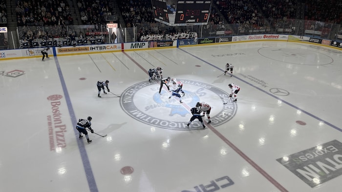 Cette photographie en plongée montre le début d'un match de hockey au Centre Georges-Vézina de Saguenay. Deux équipes, vêtues de bleu et de rouge/blanc, se font face sur la patinoire pour une mise au jeu devant une foule dense.