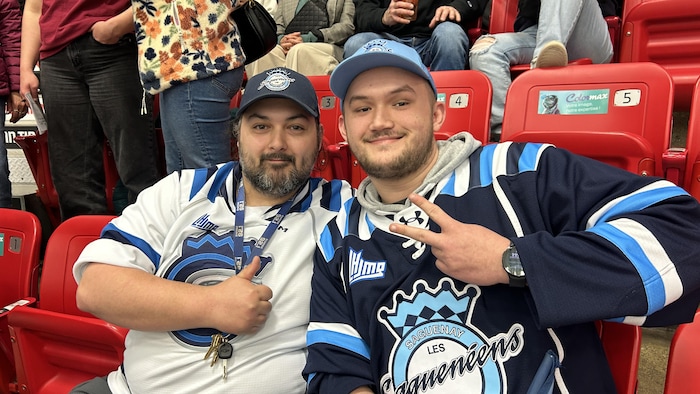 Deux hommes souriants, portant des chandails et casquettes des Saguenéens de Chicoutimi, sont assis dans des gradins rouges. L'un fait un pouce levé et l'autre un signe de la paix. Des spectateurs sont visibles en arrière-plan.