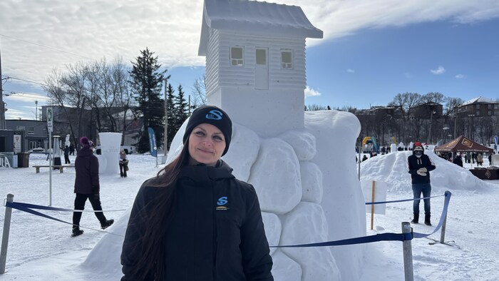 Une femme vêtue de noir pose devant une sculpture de neige représentant une petite maison sur un socle rocheux. L'événement se déroule en extérieur, sous un ciel dégagé, dans un espace public enneigé accueillant d'autres installations hivernales.