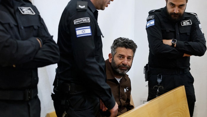 Saïf Abou Keshek est assis sur un banc dans une salle d'audience, entouré de trois membres des forces israéliennes.