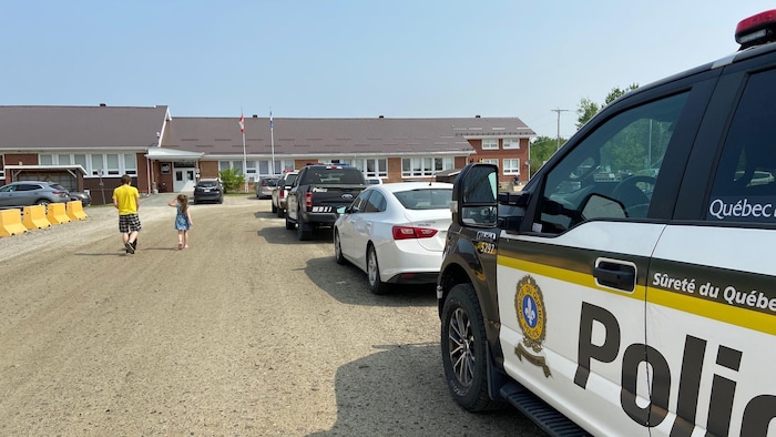 Des voitures de la Sûreté du Québec stationnées devant le centre communautaire de Saint-Lambert. Un adulte et un enfant marchent en direction de l'entrée principale.