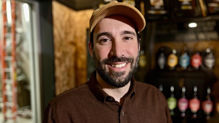 Un homme sourit à la caméra, en avant-plan de bouteilles posées sur une étagère. 