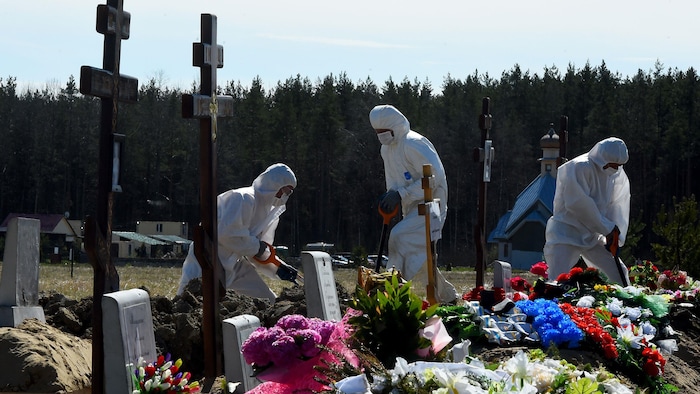 Trois travailleurs portant masque, gants et vêtement de protection creusent des tombes.