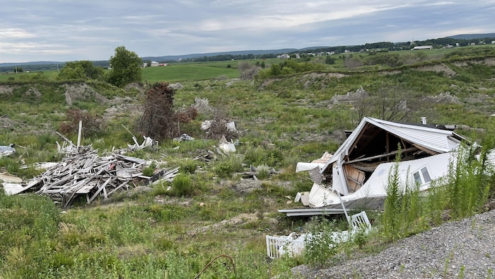 La forme d'une maison est toujours visible au travers des débris et morceaux de bois empilés au fond d'un terrain affaissé. Une petite clôture d'un ancien balcon est seule, près des débris.