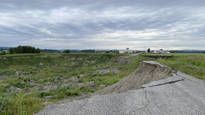 Une route d'asphalte est brisée et se transforme en un immense trou de gazon et de roche. Au loin se trouvent une maison et des bâtiments de ferme.