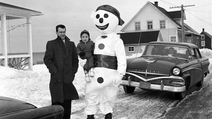 Devant une voiture typique des années 1950, un bonhomme de neige tient un enfant dans ses bras, sur une route enneigée.