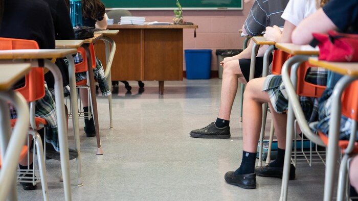 Des élèves du secondaire vus de dos dans une classe à la hauteur des jambes et des pieds, de manière à ne pouvoir les identifier.