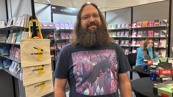 Un homme souriant à barbe longue et lunettes porte un t-shirt illustré devant un stand d'éditions.