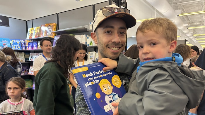 Un homme portant une casquette tient un jeune enfant dans ses bras tout en montrant un livre pour enfants.