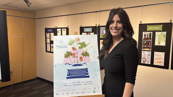 Mélanie Devost, souriante à côté de l'affiche du Salon du livre de la Côte-Nord.