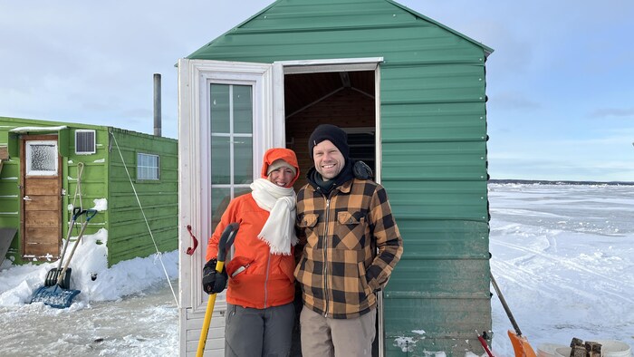Deux personnes se tiennent par l'épaule en souriant devant une petite cabane de tôle sur la glace.
