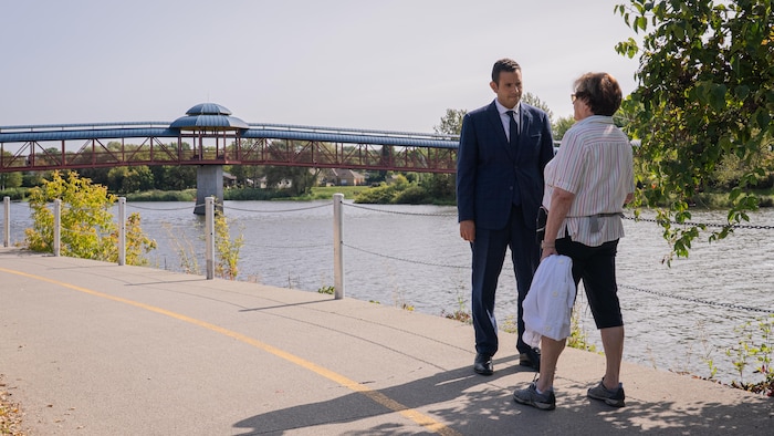 Un homme parle à une femme sur une piste cyclable devant une passerelle sur une rivière.