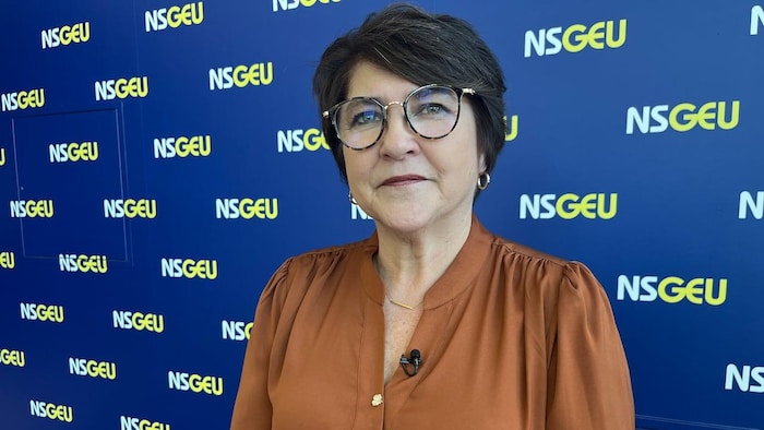 Une femme aux cheveux foncés courts, portant des lunettes, debout devant un mur bleu royal décoré de multiples logos de NSGEU.